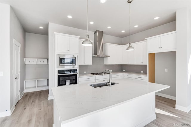 kitchen featuring wall chimney range hood, a kitchen island with sink, pendant lighting, appliances with stainless steel finishes, and white cabinetry