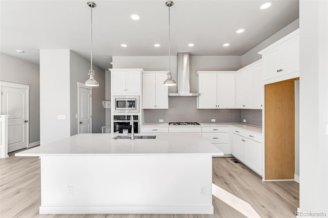 kitchen featuring wall chimney exhaust hood, pendant lighting, a center island with sink, stainless steel appliances, and white cabinets