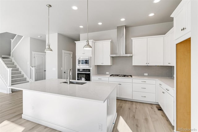 kitchen with wall chimney range hood, decorative light fixtures, a center island with sink, stainless steel appliances, and white cabinetry