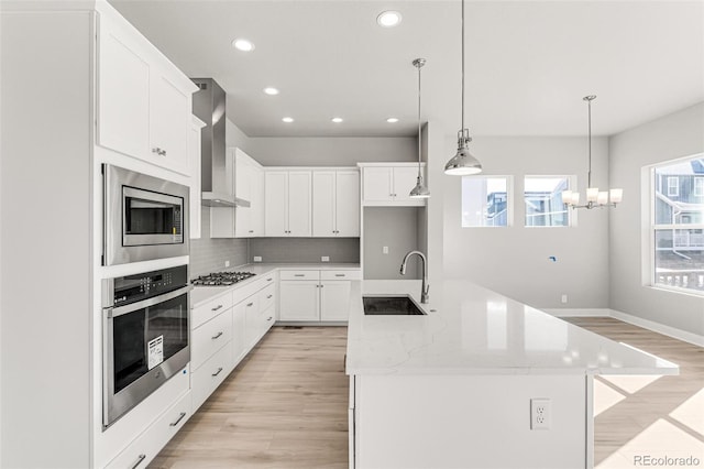 kitchen featuring wall chimney exhaust hood, a kitchen island with sink, sink, stainless steel appliances, and white cabinetry