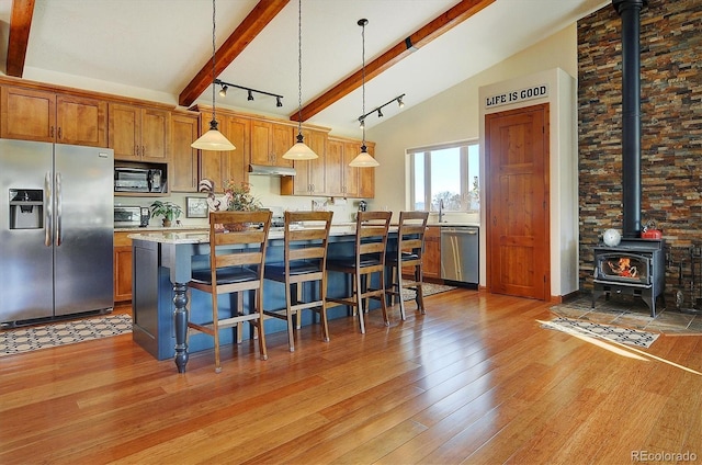 kitchen featuring under cabinet range hood, stainless steel appliances, light wood-style floors, beam ceiling, and a wood stove