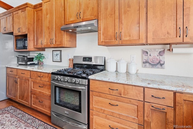 kitchen with black microwave, stainless steel gas range oven, light stone counters, under cabinet range hood, and brown cabinetry