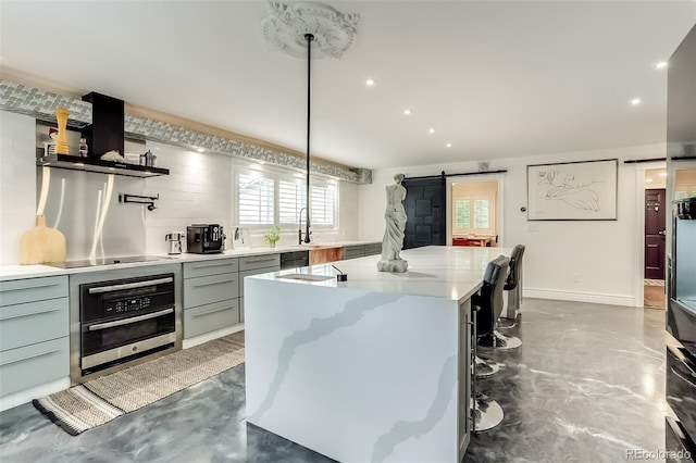 kitchen with a barn door, wall chimney range hood, stainless steel oven, black electric stovetop, and a kitchen island