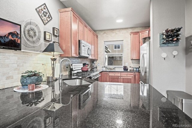 kitchen featuring brown cabinetry, backsplash, a sink, dark stone countertops, and stainless steel appliances