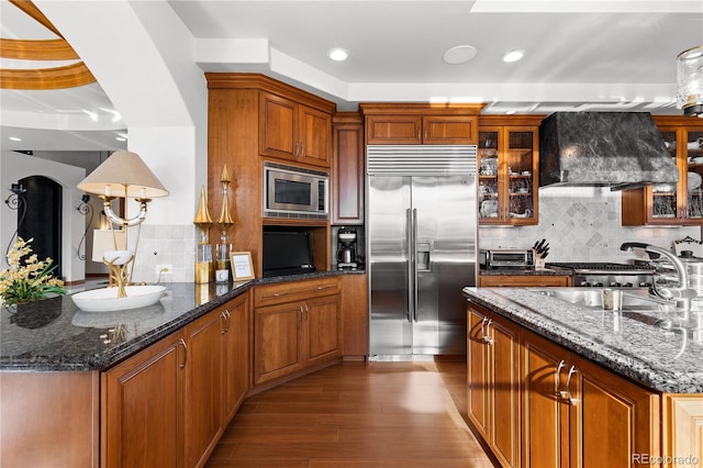 kitchen with wall chimney exhaust hood, sink, backsplash, dark stone countertops, and built in appliances