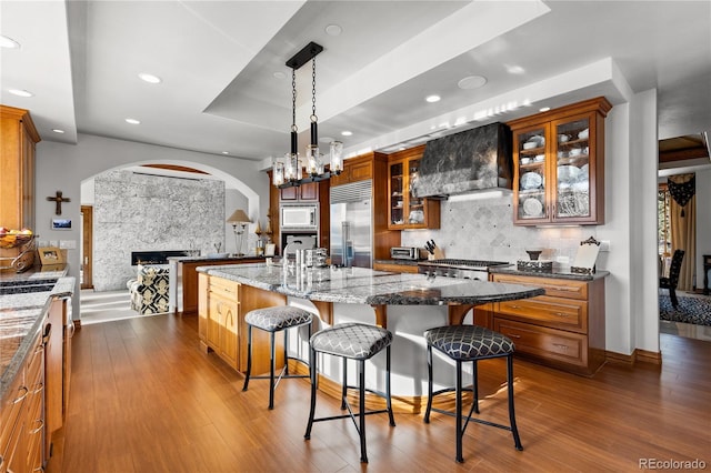 kitchen with pendant lighting, a kitchen island with sink, a breakfast bar, wall chimney range hood, and built in appliances