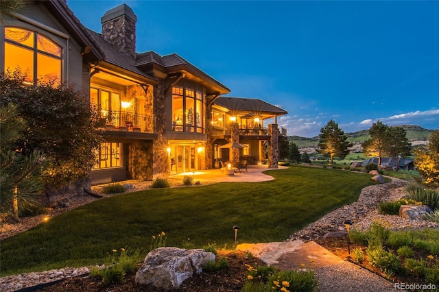 back house at dusk with a balcony, a patio area, and a yard