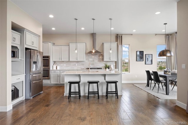 kitchen featuring wall chimney range hood, appliances with stainless steel finishes, tasteful backsplash, an island with sink, and pendant lighting