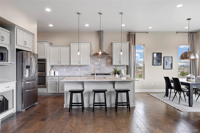 kitchen featuring appliances with stainless steel finishes, wall chimney exhaust hood, backsplash, a breakfast bar, and a kitchen island with sink