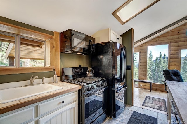 kitchen with tile countertops, sink, black appliances, vaulted ceiling with skylight, and white cabinetry