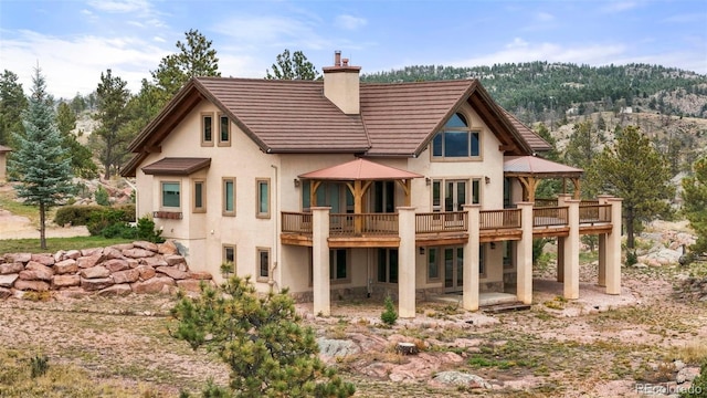 rear view of property with a forest view, a chimney, a wooden deck, a patio area, and stucco siding