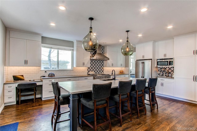 kitchen with wall chimney range hood, appliances with stainless steel finishes, a breakfast bar, and dark wood-style flooring