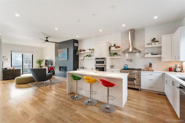 kitchen featuring a tile fireplace, wall chimney exhaust hood, appliances with stainless steel finishes, a breakfast bar, and open shelves