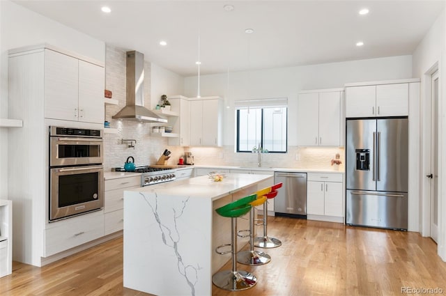 kitchen featuring stainless steel appliances, a breakfast bar, light wood-style floors, wall chimney range hood, and tasteful backsplash