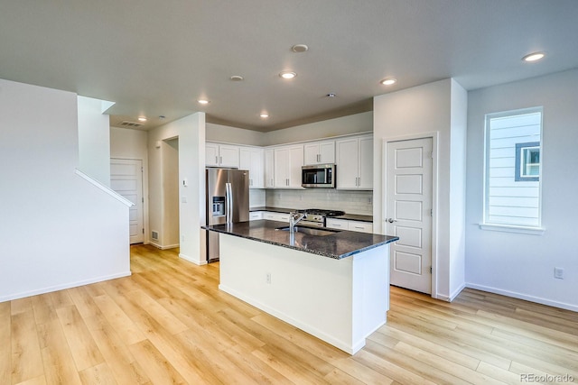 kitchen featuring appliances with stainless steel finishes, dark stone countertops, white cabinets, backsplash, and a kitchen island with sink