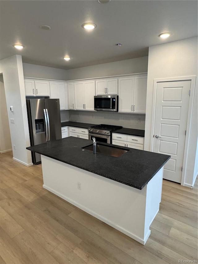 kitchen with appliances with stainless steel finishes, white cabinetry, backsplash, light hardwood / wood-style floors, and a center island with sink