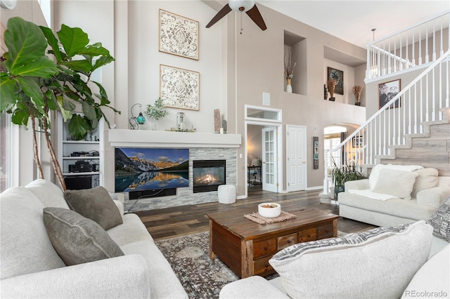 living room with ceiling fan, a stone fireplace, dark hardwood / wood-style flooring, and a towering ceiling