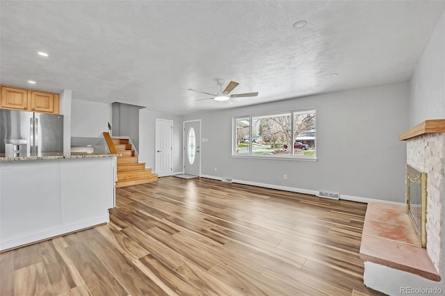 unfurnished living room with ceiling fan, light wood-style flooring, a textured ceiling, a glass covered fireplace, and stairs