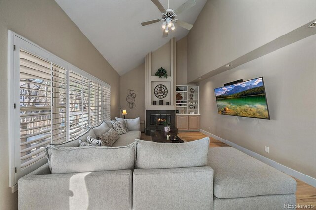 living area with a large fireplace, ceiling fan, wood finished floors, high vaulted ceiling, and baseboards