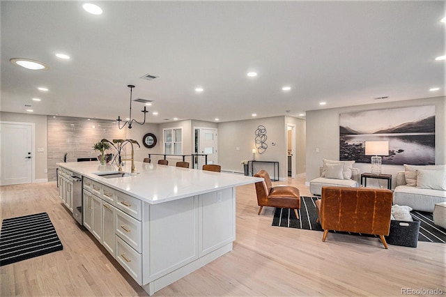 kitchen with an island with sink, sink, hanging light fixtures, stainless steel dishwasher, and light wood-type flooring