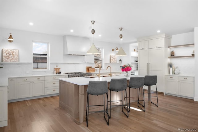 kitchen with a kitchen island with sink, sink, hanging light fixtures, light hardwood / wood-style floors, and custom range hood