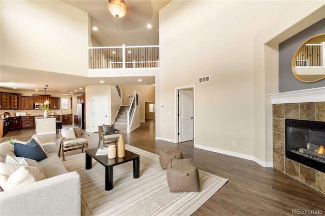 living room with visible vents, a tiled fireplace, stairway, baseboards, and dark wood-style flooring