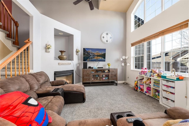 carpeted living room with stairway, a towering ceiling, a glass covered fireplace, ceiling fan, and baseboards