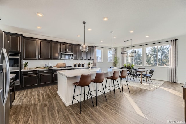 kitchen featuring wood finished floors, an island with sink, stainless steel appliances, light countertops, and a kitchen breakfast bar