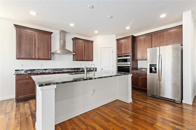 kitchen with an island with sink, dark wood-type flooring, appliances with stainless steel finishes, and wall chimney range hood