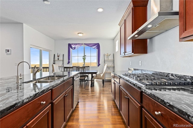 kitchen featuring a textured ceiling, light hardwood / wood-style flooring, wall chimney range hood, appliances with stainless steel finishes, and dark stone countertops