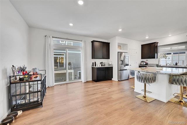 kitchen with dark brown cabinetry, a breakfast bar, stainless steel refrigerator with ice dispenser, light stone countertops, and light hardwood / wood-style flooring