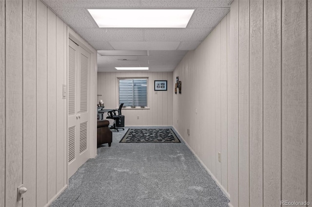 hallway featuring carpet floors, a desk, wood walls, a paneled ceiling, and baseboards