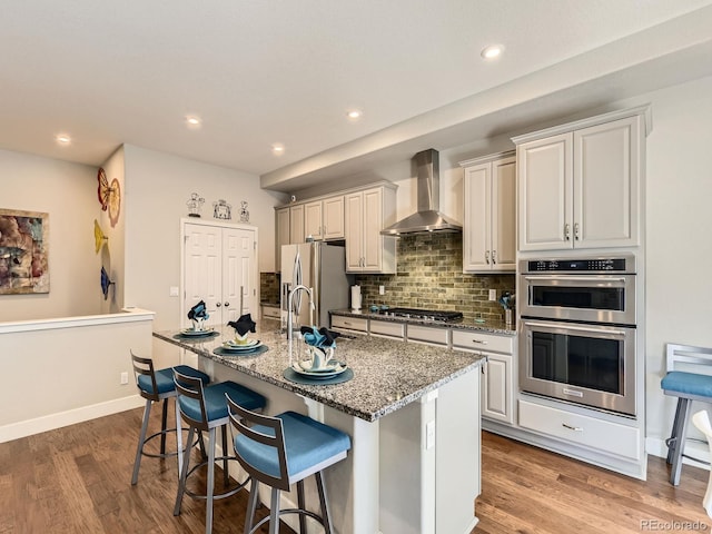 kitchen with stainless steel appliances, a sink, wall chimney range hood, backsplash, and a center island with sink