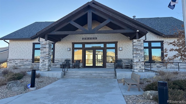 entrance to property with stone siding, french doors, roof with shingles, and stucco siding