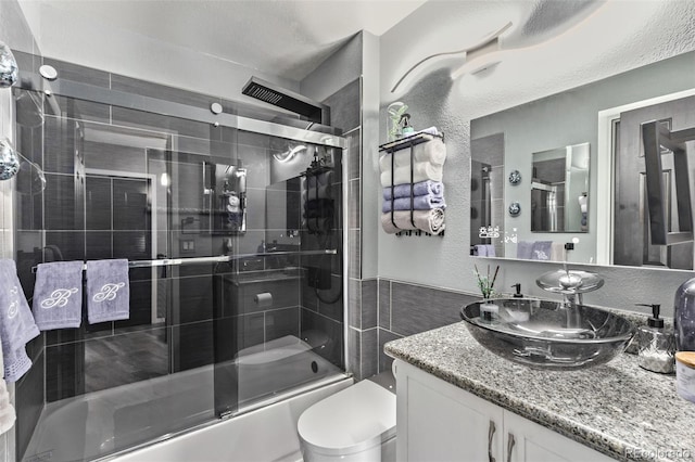 full bathroom featuring a textured wall, bath / shower combo with glass door, vanity, and toilet