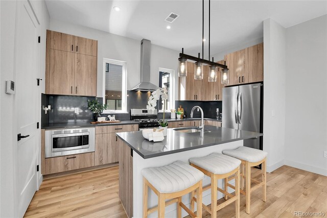 kitchen featuring a breakfast bar, a sink, appliances with stainless steel finishes, dark countertops, and wall chimney range hood