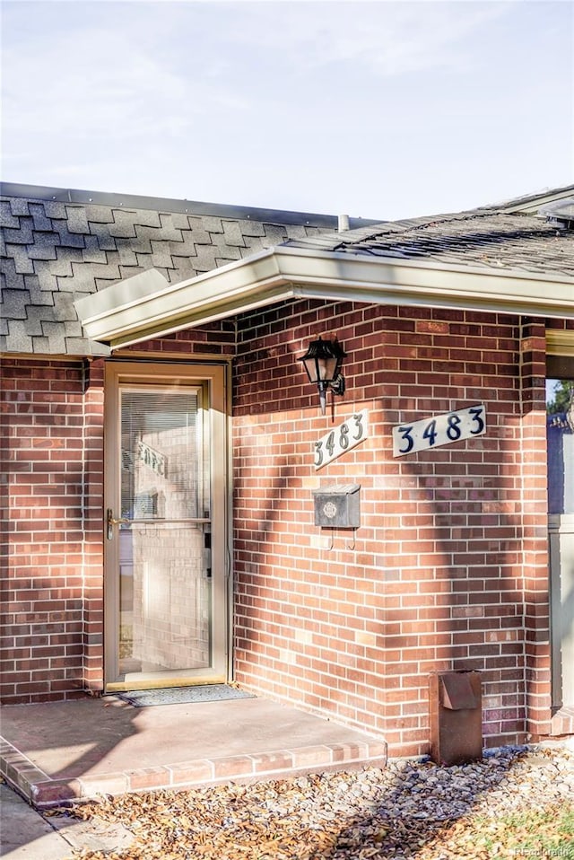 view of exterior entry with brick siding and a shingled roof