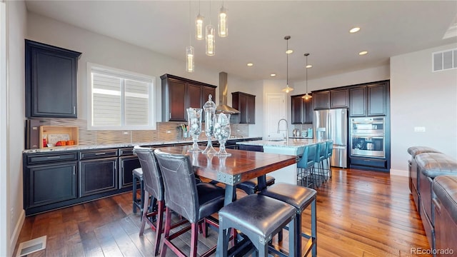 kitchen with stainless steel appliances, a kitchen island with sink, visible vents, and wall chimney range hood