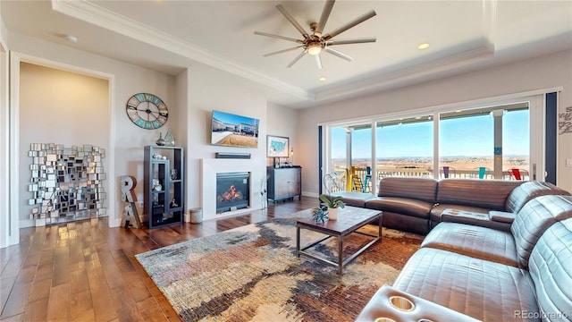 living area with baseboards, a raised ceiling, wood-type flooring, a glass covered fireplace, and crown molding