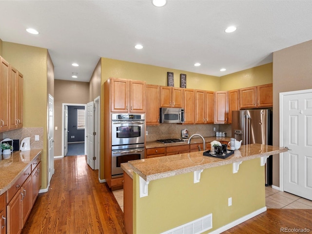 3853 S Malta St, Aurora, CO 80013 - kitchen featuring stainless steel appliances, a kitchen breakfast bar, a center island with sink, tasteful backsplash, and light wood-type flooring