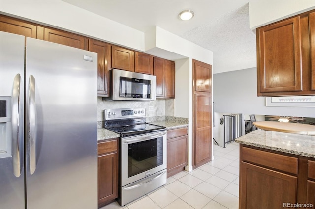 kitchen with stainless steel appliances, backsplash, light tile patterned floors, light stone counters, and brown cabinets