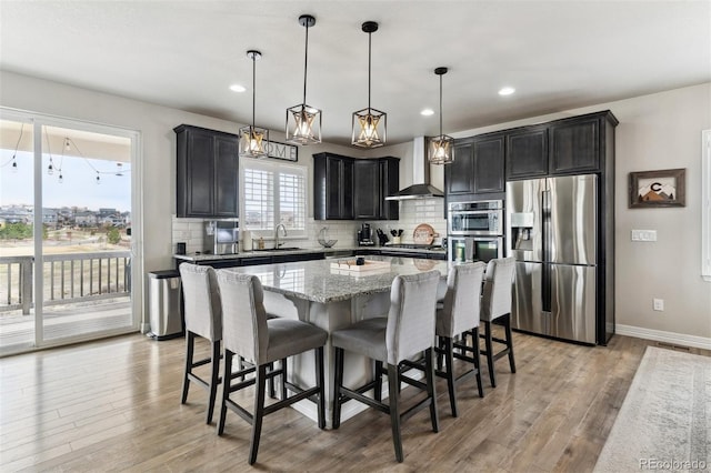 kitchen featuring decorative backsplash, stainless steel appliances, a kitchen island, light stone counters, and wall chimney range hood