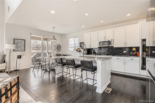 kitchen with decorative light fixtures, stainless steel appliances, white cabinets, an island with sink, and light stone countertops