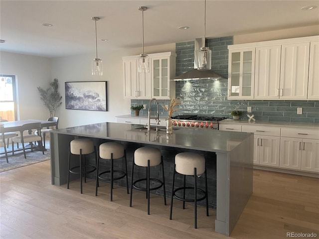 kitchen with light wood-type flooring, a center island with sink, a sink, tasteful backsplash, and wall chimney exhaust hood