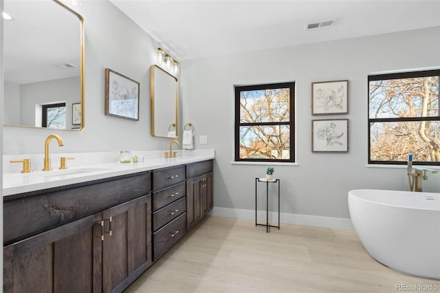 bathroom featuring wood finished floors, a sink, double vanity, a freestanding bath, and visible vents
