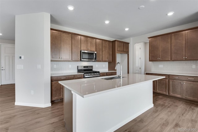 kitchen with stainless steel appliances, light wood-type flooring, sink, backsplash, and a kitchen island with sink