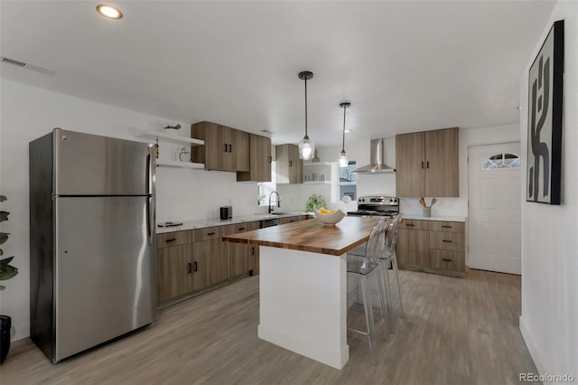 kitchen with wall chimney exhaust hood, a center island, decorative light fixtures, butcher block counters, and appliances with stainless steel finishes