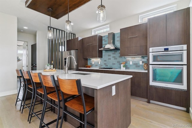 kitchen with pendant lighting, a breakfast bar, wall chimney range hood, sink, and an island with sink