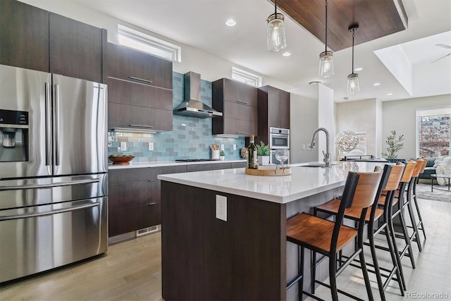 kitchen featuring pendant lighting, a center island with sink, wall chimney range hood, sink, and appliances with stainless steel finishes