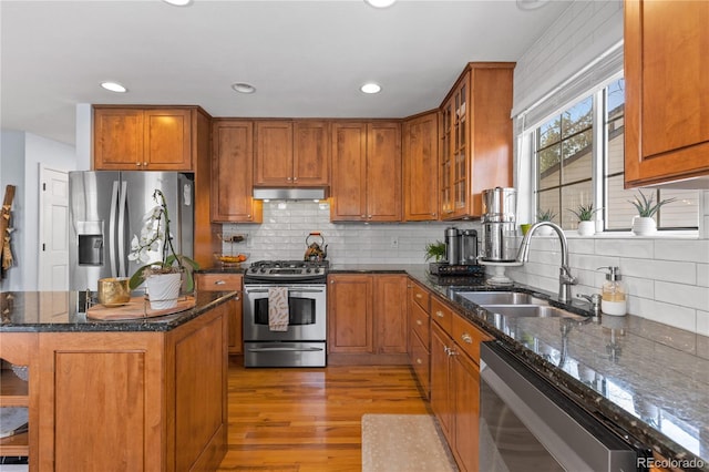 kitchen featuring under cabinet range hood, a sink, dark stone counters, appliances with stainless steel finishes, and brown cabinetry